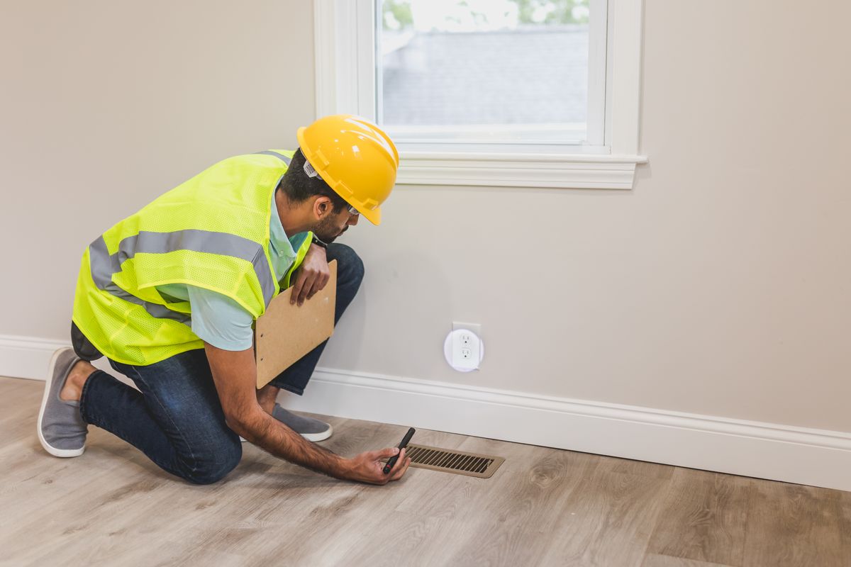 Inspector in a yellow hard hat and safety vest kneeling on a wood-look floor, examining a metal floor vent with a flashlight